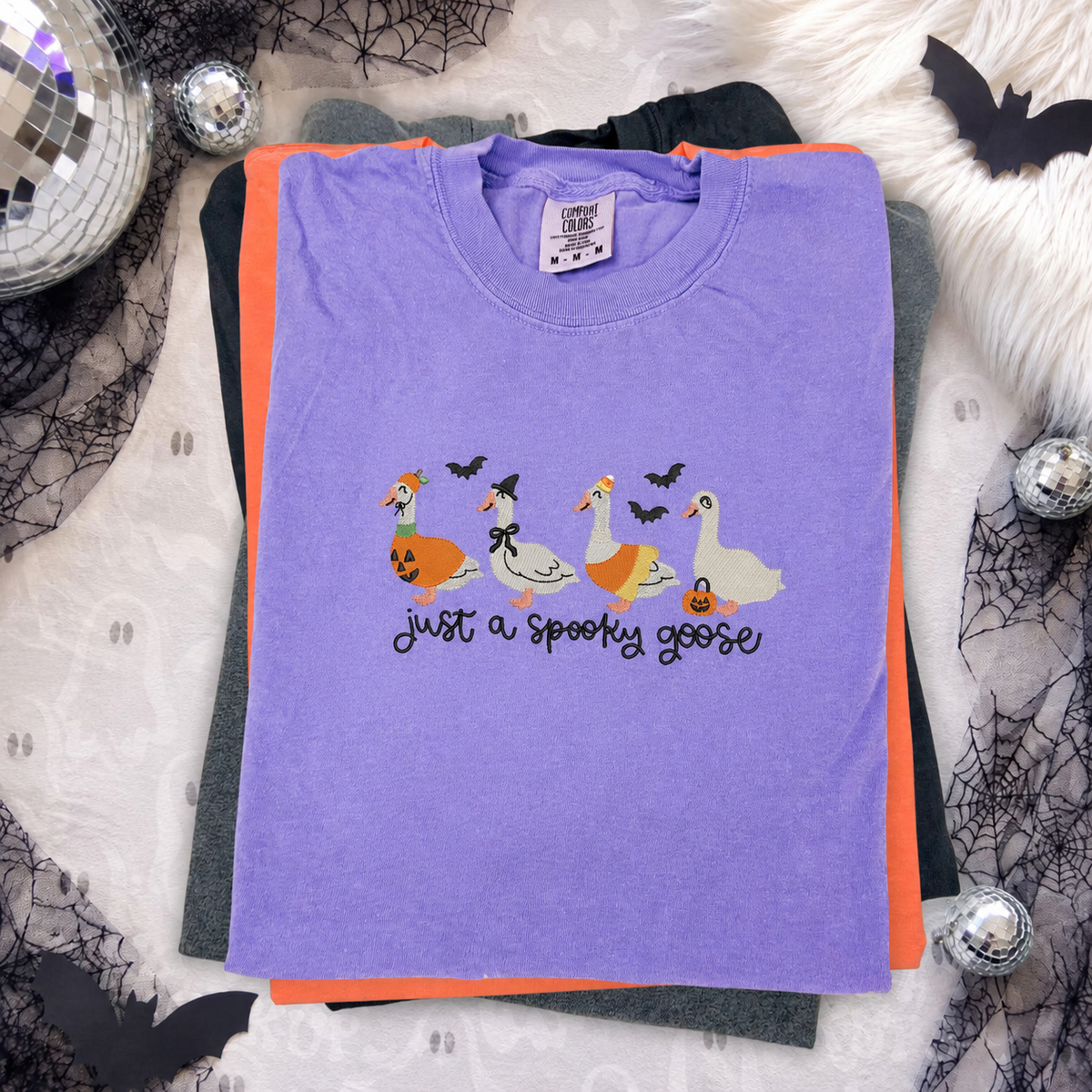 purple comfort color t-shirt with a 'just a spooky goose' embroidered design featuring four halloween costume decorated geese and bats, 