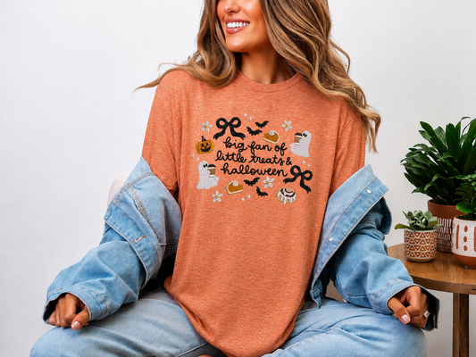 A woman wearing a heathered orange crewneck t-shirt with a embroidered Halloween graphic that reads 'big fan of little treats & halloween' with ghosts, bats, and pumpkins, bows and flowers styled with a light blue denim jacket draped off her shoulders.