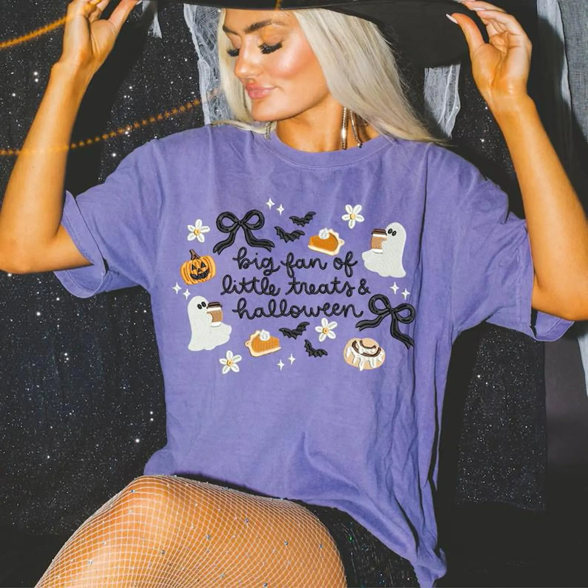 A woman wearing a purple embroidered comfort color t-shirt with a Halloween theme that reads 'big fan of little treats & halloween' surrounded by illustrations of ghosts, pumpkins, treats, bows and bats.